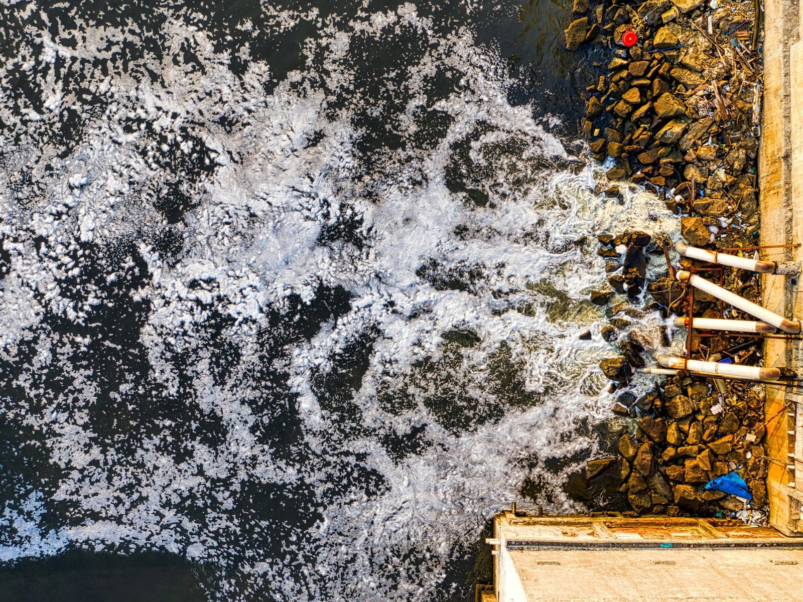An aerial view of ocean waves splashing against a rocky shore near a concrete wall in Jakarta, Indonesia.